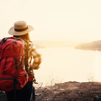 Bild einer Frau mit Trekking-Kleidung und mit grossem Rucksack, die in wunderschöner Natur auf einen See im Abendlicht schaut.