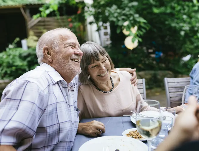 Bild eines Rentnerpaares, dass zusammen mit anderen Personen an einem Gartentisch sitzt und lachend diskutiert.